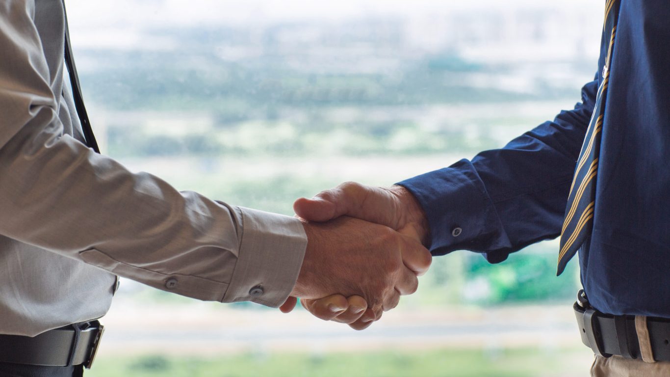 Closeup of two businessmen shaking hands with blurry city view in background. People are seen partly.